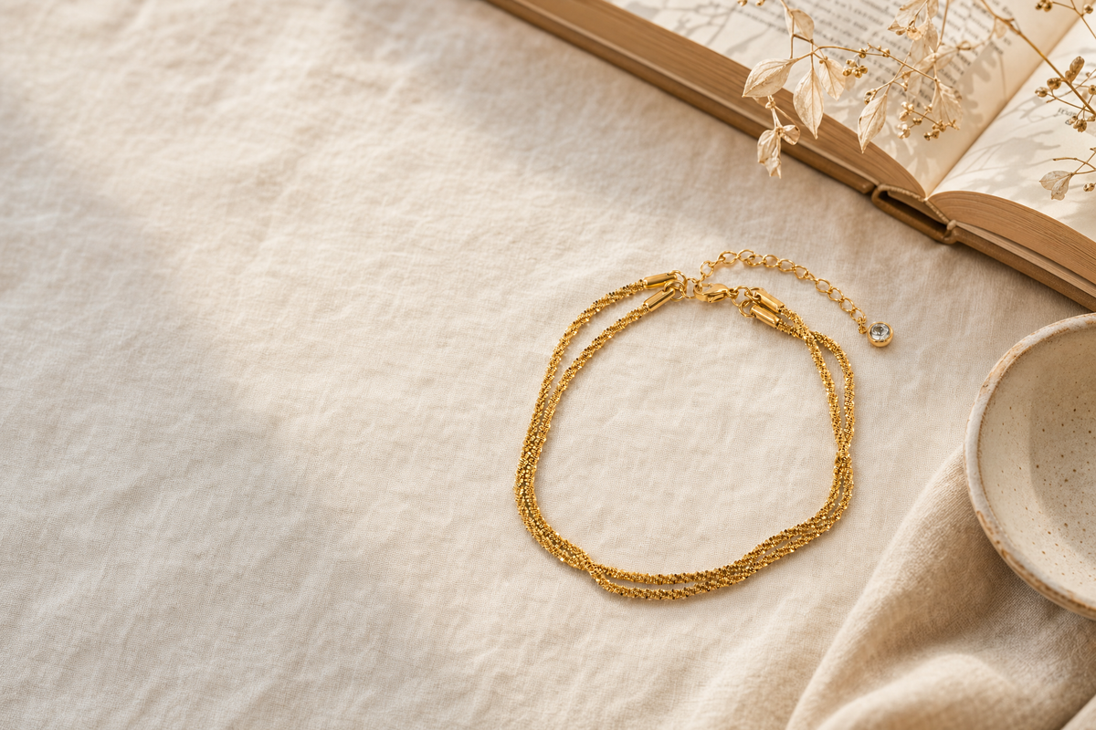 A gold-tone multi-strand anklet with a lobster clasp and extender chain laid flat on cream linen beside an open book