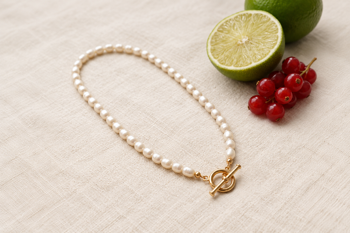 A white pearl necklace with a gold-tone toggle clasp rests on warm cream linen, with a halved lime