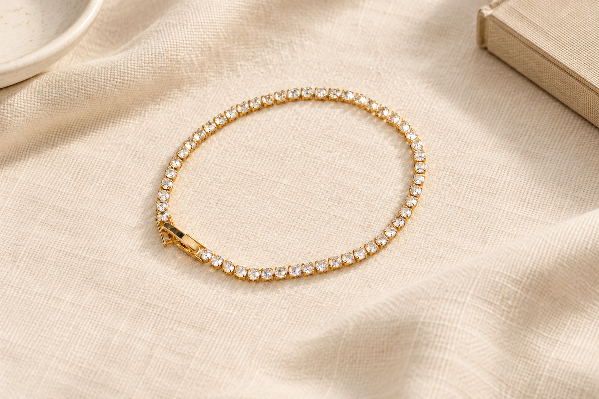 A gold-tone tennis bracelet with a single row of clear round stones and a rectangular clasp rests on cream linen in warm natural light, framed by a partial ceramic dish and a linen-covered book.