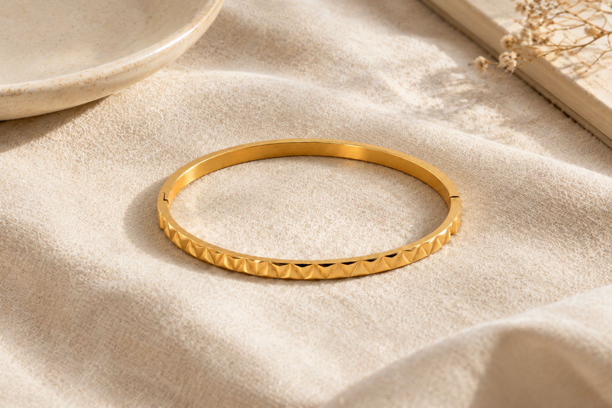 A gold-colored oval bangle with repeating triangular facets along the front rests on cream linen in warm natural light, with a partial ceramic dish at the upper left and a book with dried flowers at the upper right.