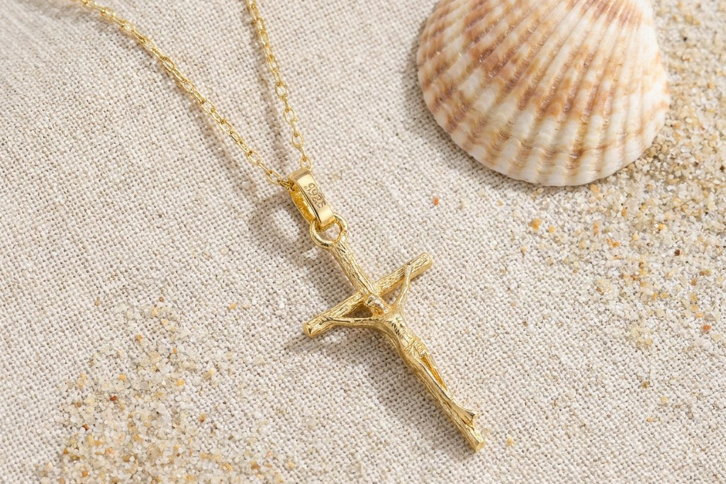 Gold-colored crucifix pendant and fine chain on cream linen, with scattered sand and a seashell in soft natural light.