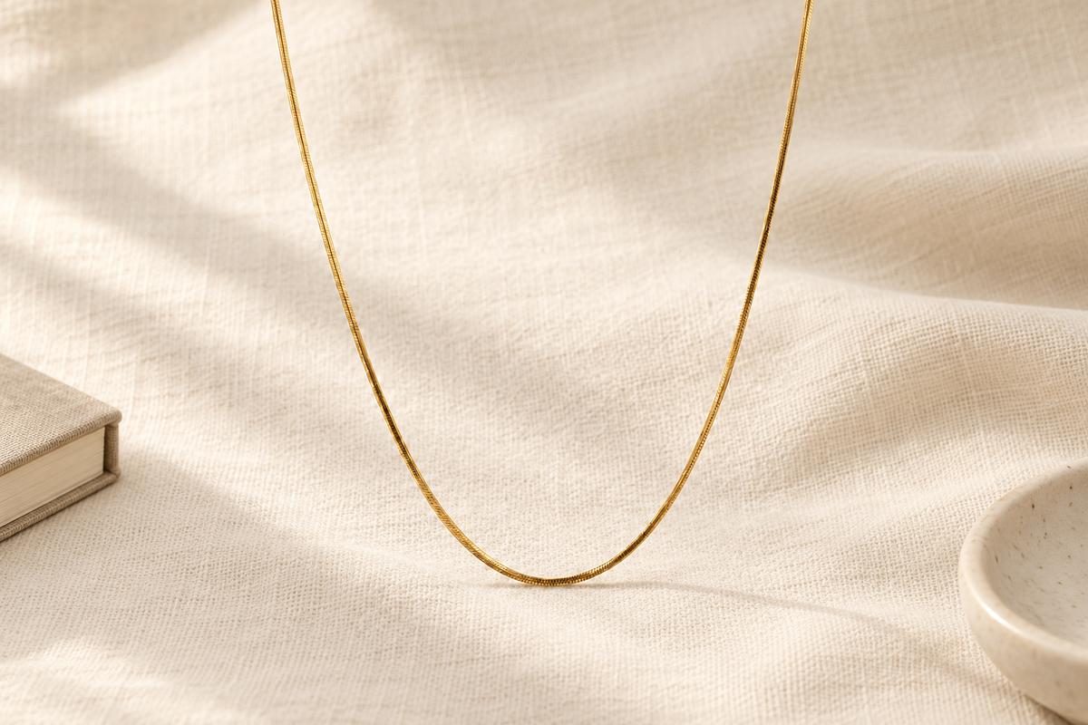 A fine gold chain forms a long U shape against warm cream linen in soft natural light