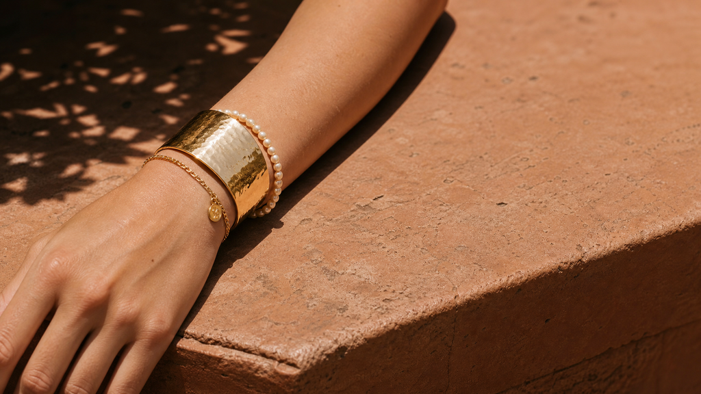 A gold cuff, chain bracelet and pearl strand stacked on a wrist in sunlight