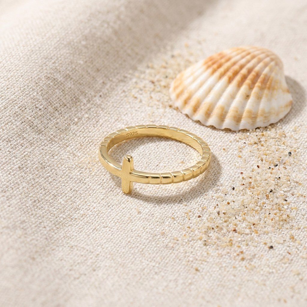 Gold Plated Cross Ring in Stainless Steel on raw linen beside a scallop shell and a scatter of fine sand