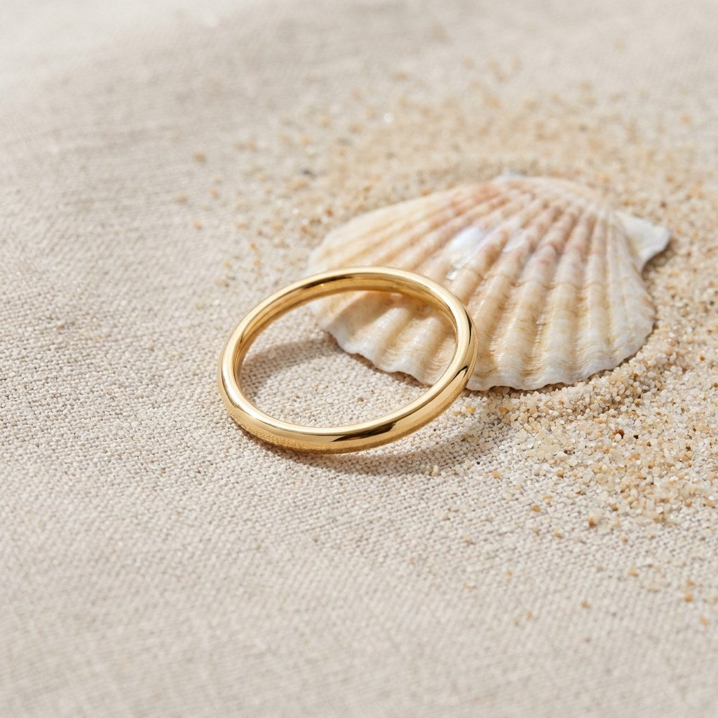 2mm Gold Stacker Band Ring on raw linen beside a scallop shell and a scatter of fine sand
