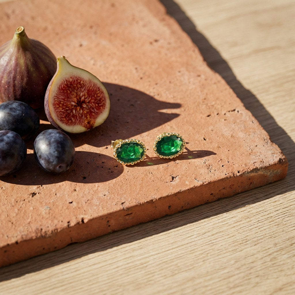 Emerald Oval Stud Earrings on a weathered terracotta tile with a cut fig and dark plums