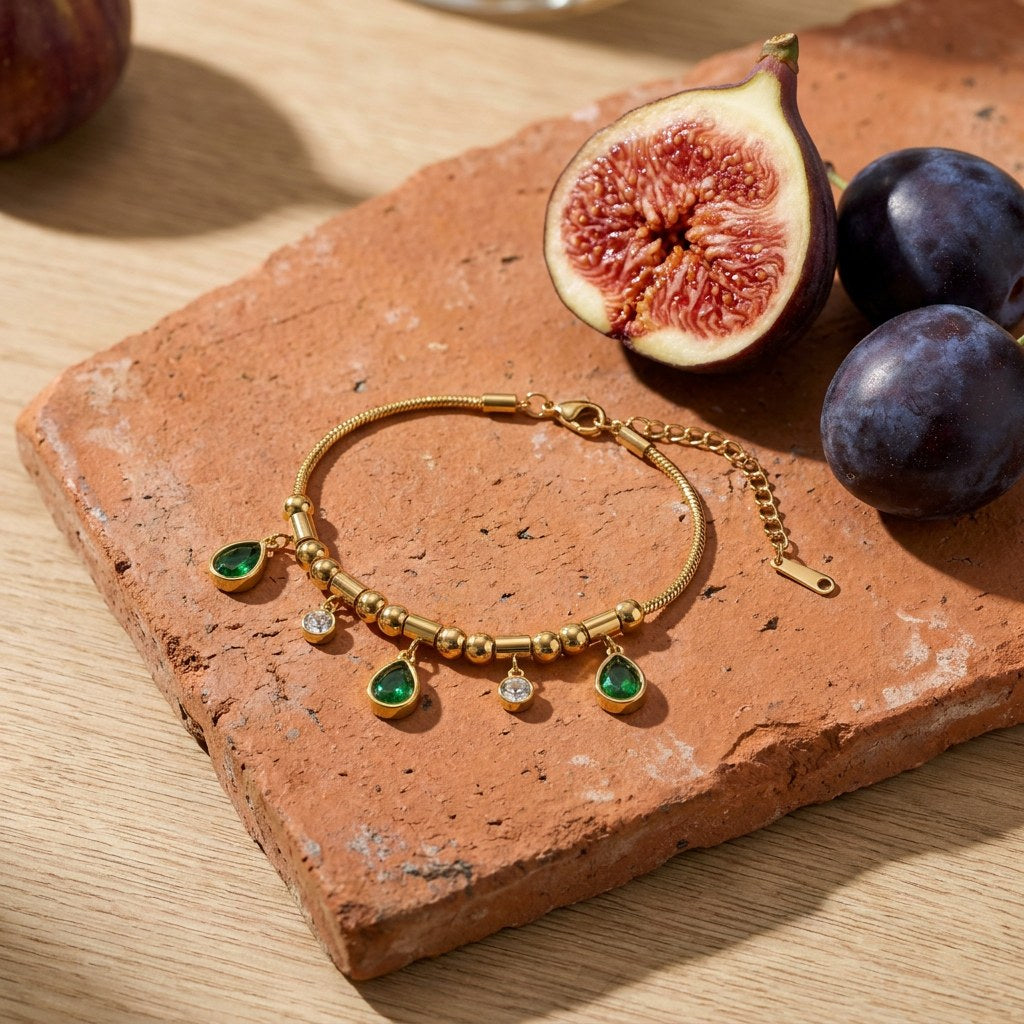 Emerald Teardrop Charm Bracelet on a weathered terracotta tile with a cut fig and dark plums