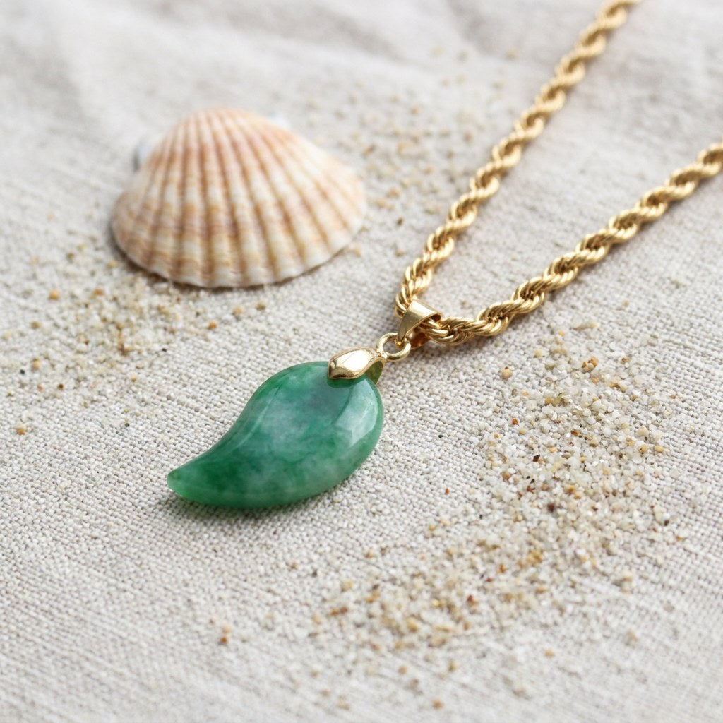 Gold Jade Comma Pendant Necklace on raw linen beside a scallop shell and a scatter of fine sand