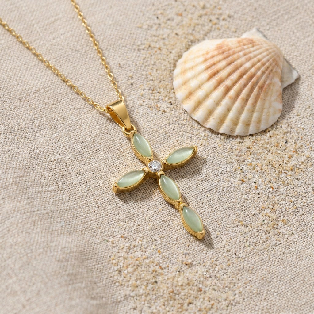 Green Cat's Eye Cross Necklace on raw linen beside a scallop shell and a scatter of fine sand