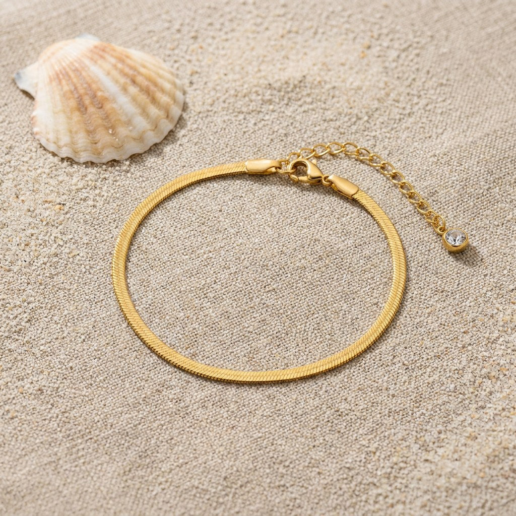 Herringbone Anklet on raw linen beside a scallop shell and a scatter of fine sand