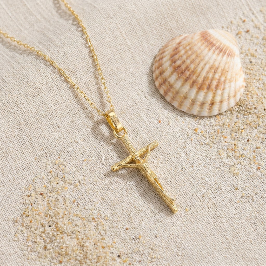 Hypoallergenic 14k Gold Plated Crucifix Pendant Necklace on raw linen beside a scallop shell and a scatter of fine sand