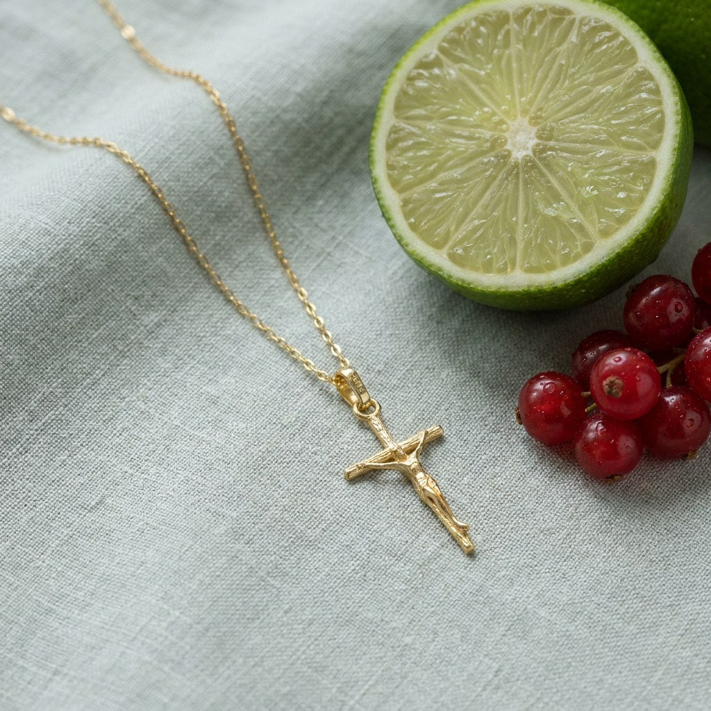 Hypoallergenic 14k Gold Plated Crucifix Pendant Necklace on pale grey-green linen beside a halved lime and red currants