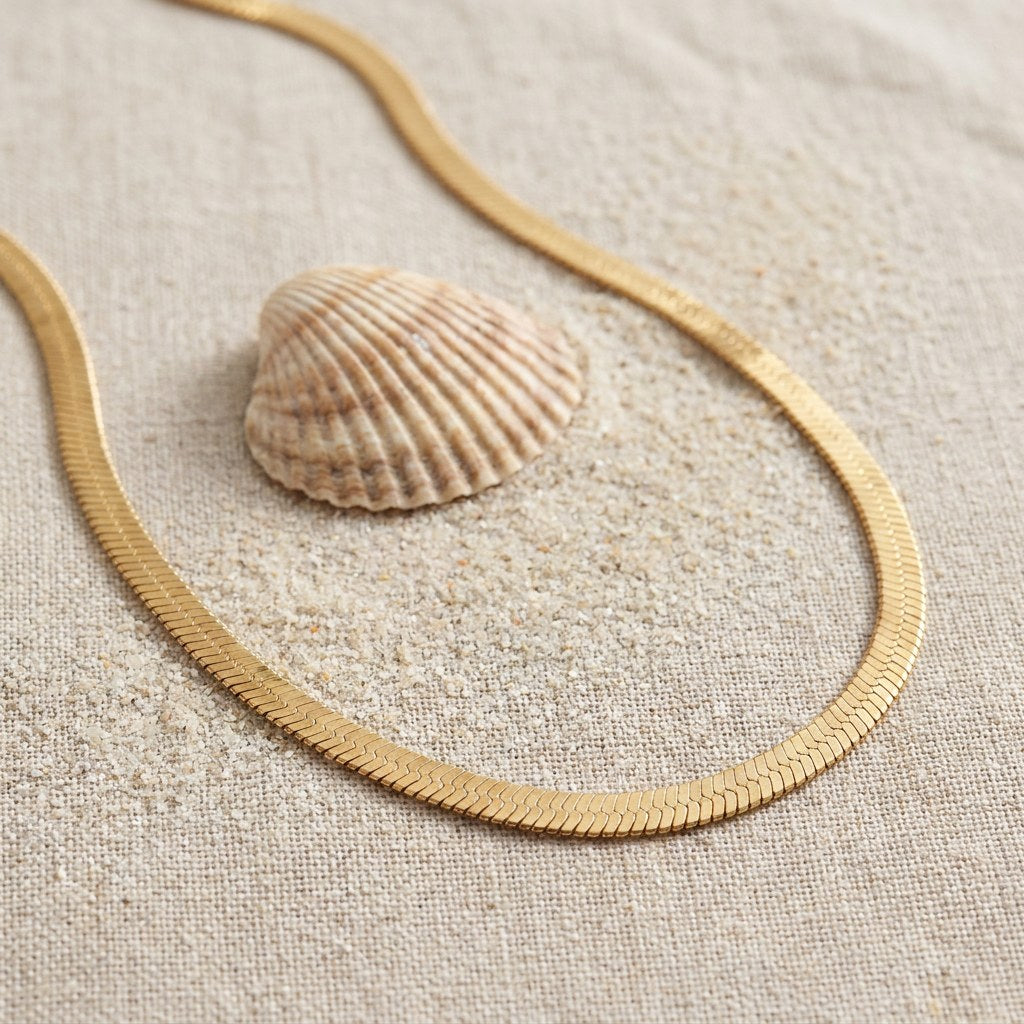 Mata Snake Chain Necklace on raw linen beside a scallop shell and a scatter of fine sand