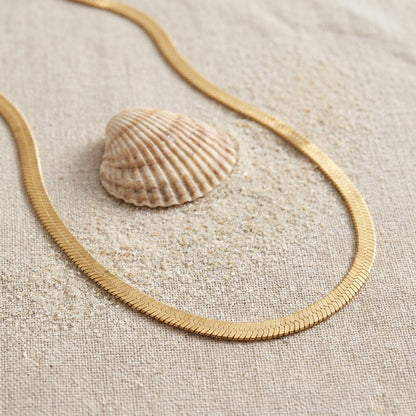 Mata Snake Chain Necklace on raw linen beside a scallop shell and a scatter of fine sand