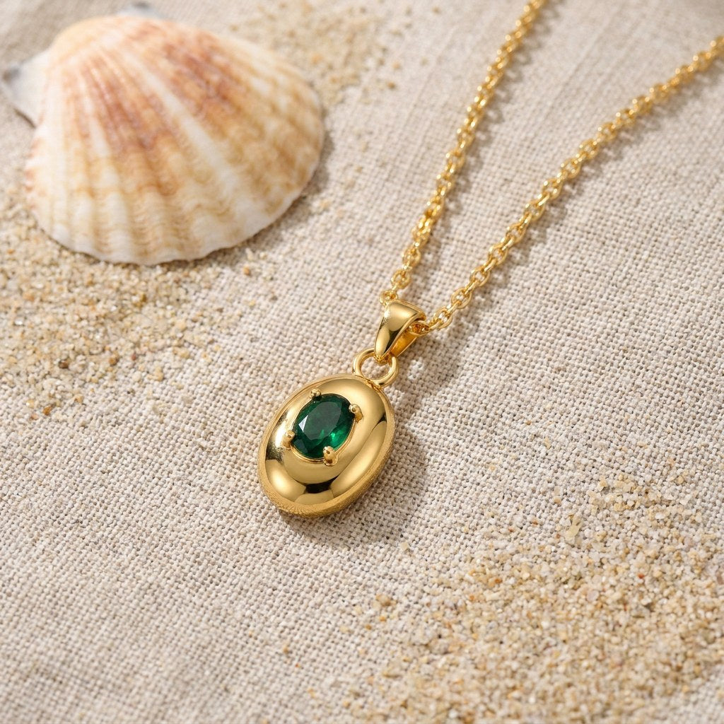 Oval Pendant Emerald on raw linen beside a scallop shell and a scatter of fine sand