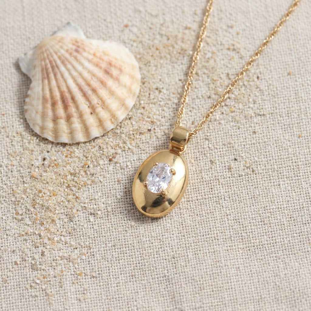 Oval Pendant Necklace on raw linen beside a scallop shell and a scatter of fine sand