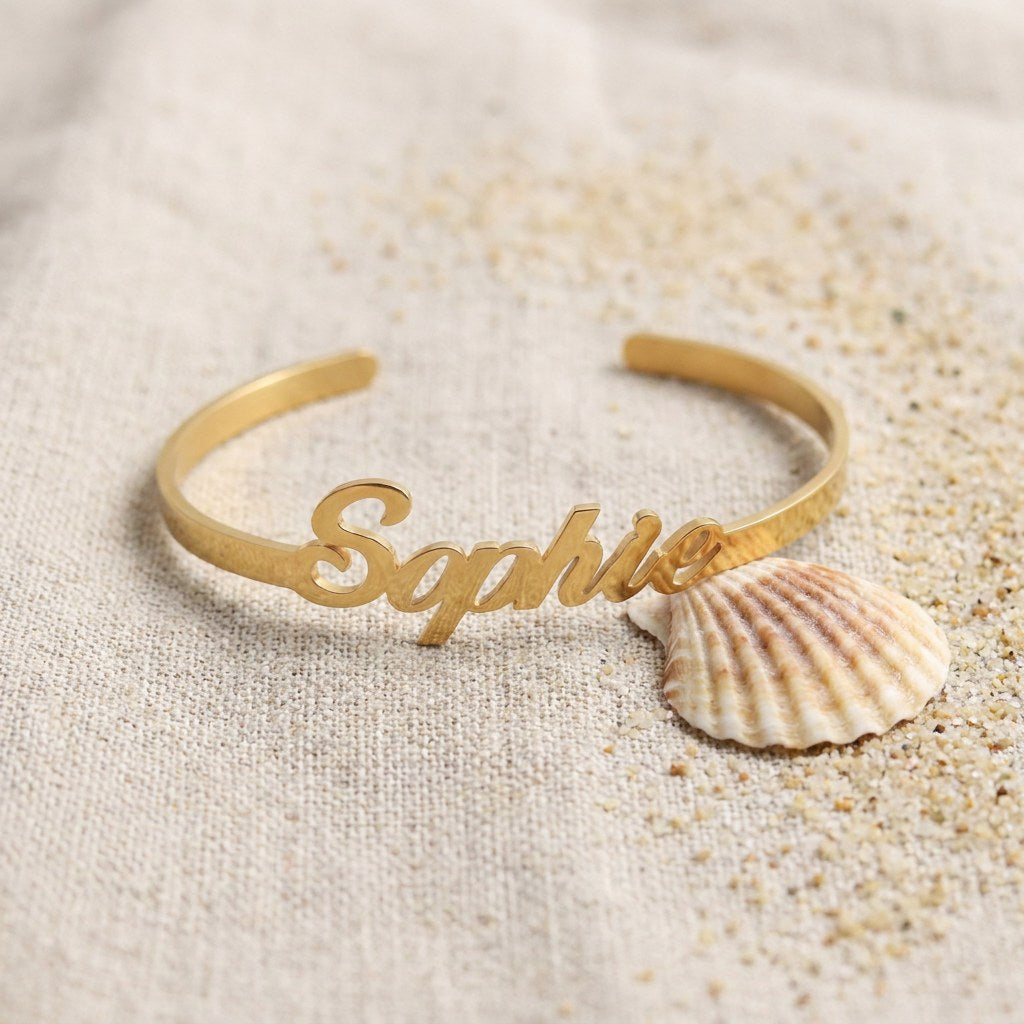Personalized Gold Name Cuff Bracelet on raw linen beside a scallop shell and a scatter of fine sand
