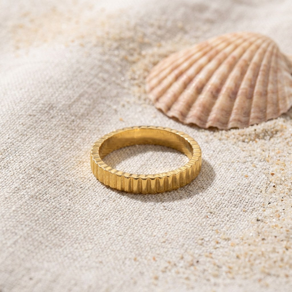 Ribbed Thin Band Ring on raw linen beside a scallop shell and a scatter of fine sand