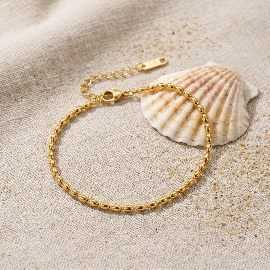 Rice Bead Anklet on raw linen beside a scallop shell and a scatter of fine sand