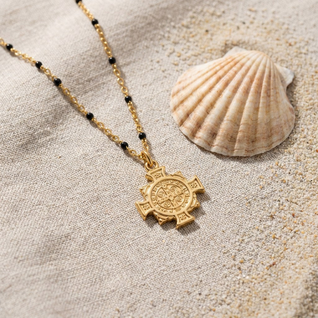 Saint Benedict Medal Necklace on raw linen beside a scallop shell and a scatter of fine sand