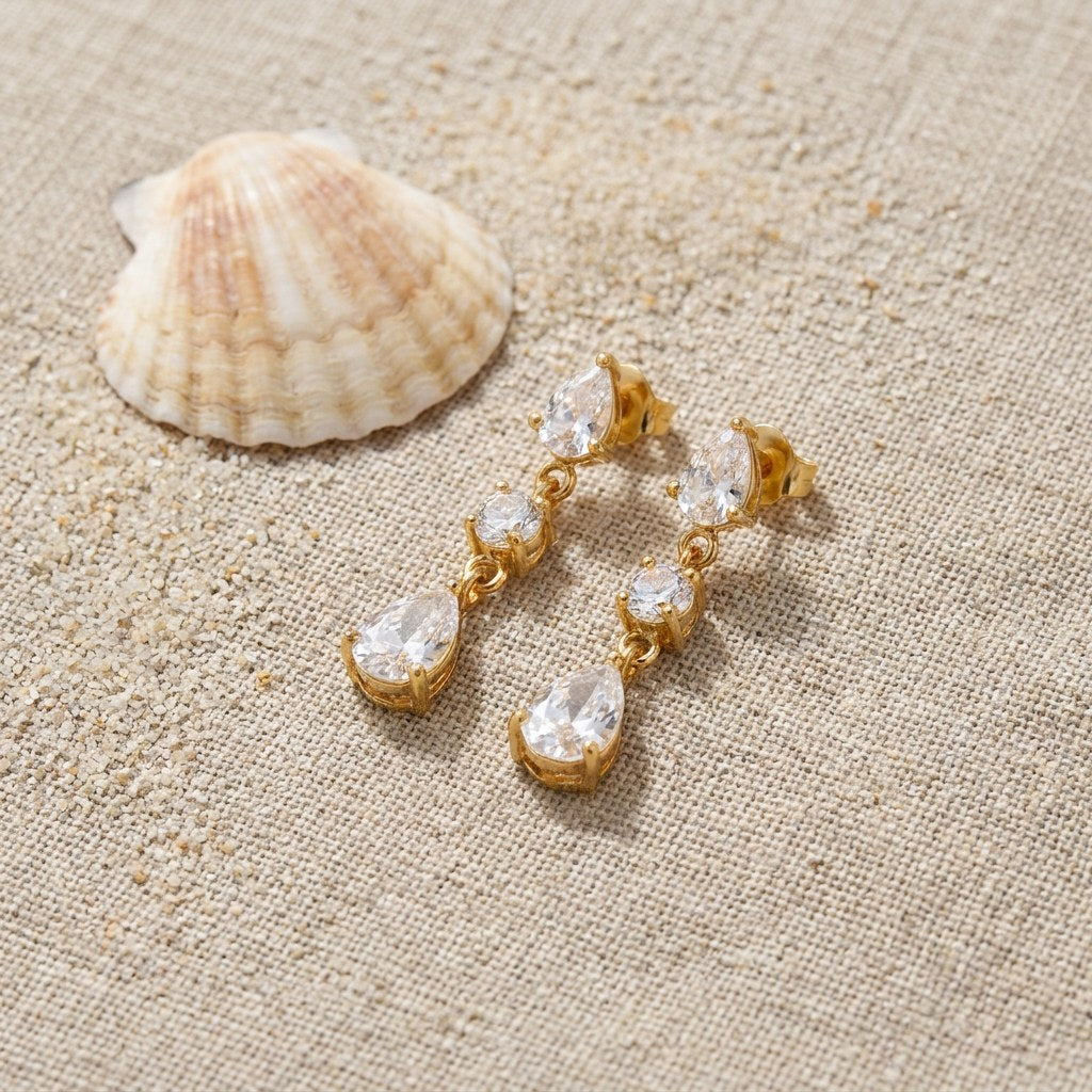 Triple CZ Drop Earrings on raw linen beside a scallop shell and a scatter of fine sand