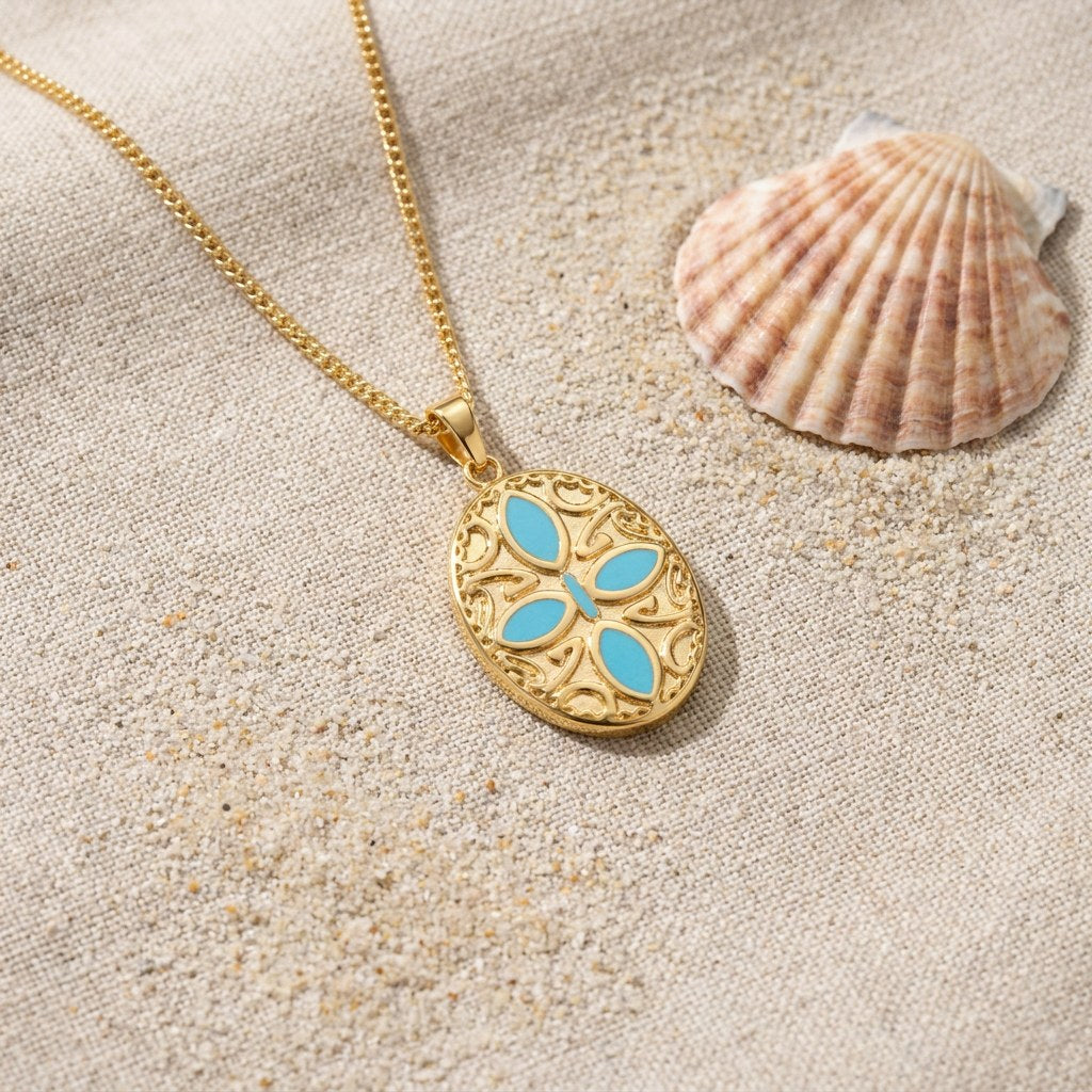 Oval Blue Petals Pendant Necklace on raw linen beside a scallop shell and a scatter of fine sand