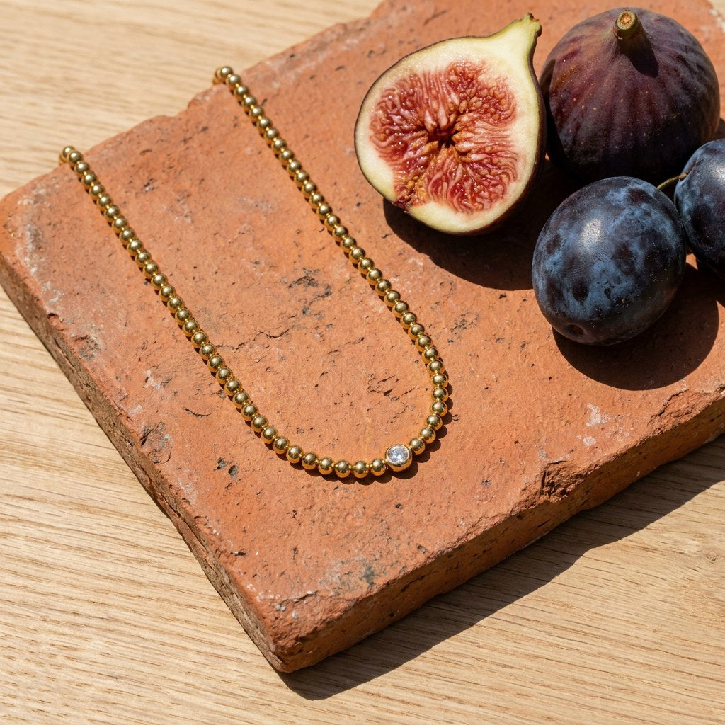 Dainty Solitaire Beaded Necklace on a weathered terracotta tile with a cut fig and dark plums