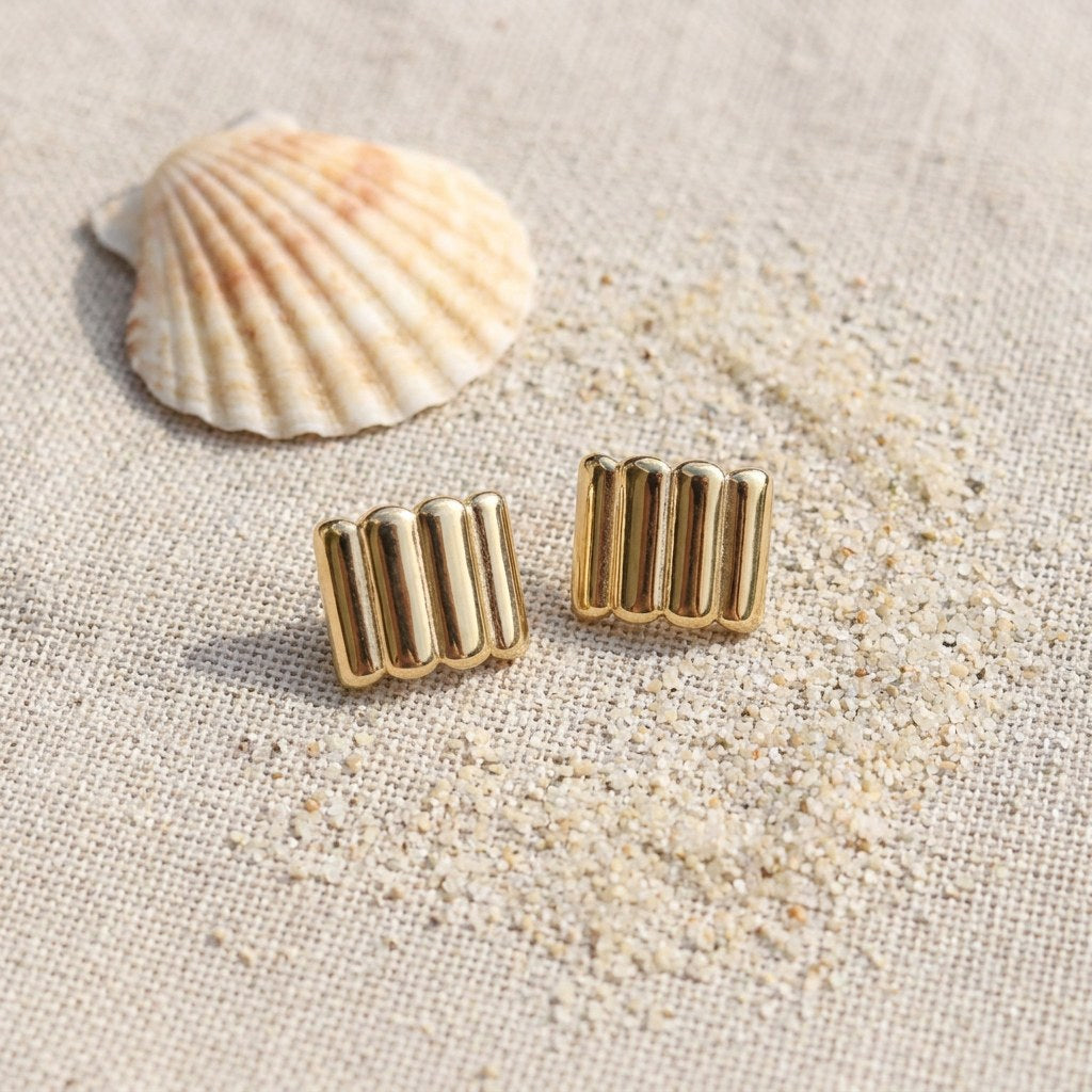 Ridged gold bar stud earrings on raw linen beside a scallop shell and a scatter of fine sand