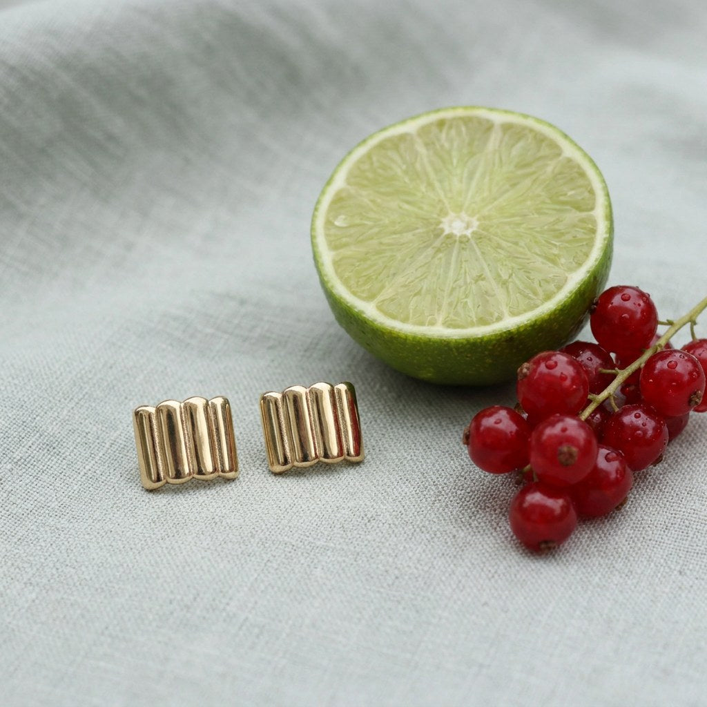 Ridged gold bar stud earrings on pale grey-green linen beside a halved lime and red currants