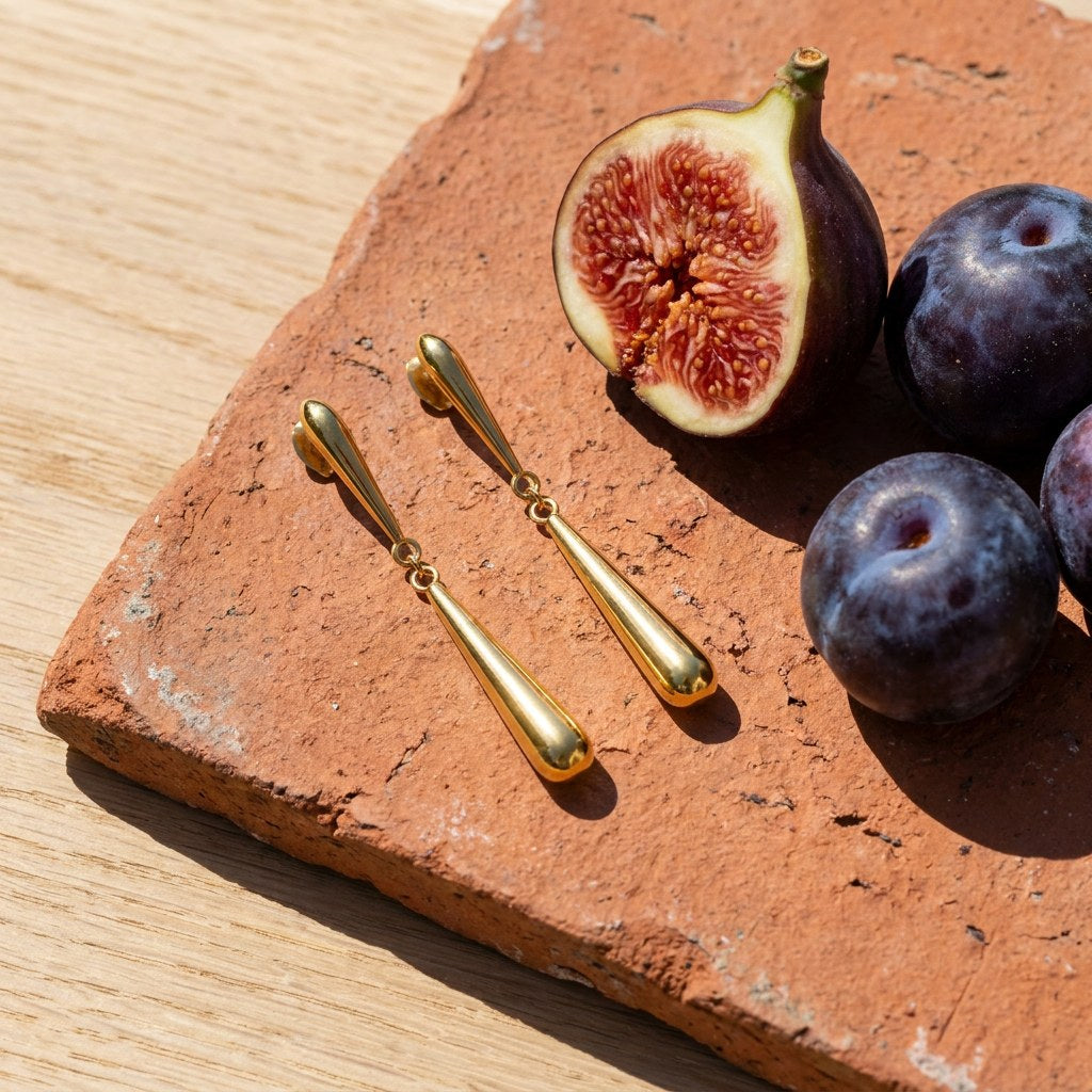 Gold Teardrop Dangle Earrings on a weathered terracotta tile with a cut fig and dark plums