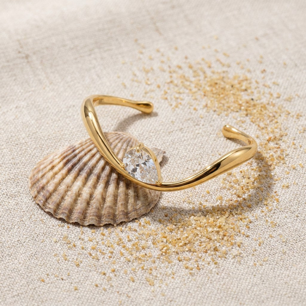 Wavy Pear Cuff on raw linen beside a scallop shell and a scatter of fine sand